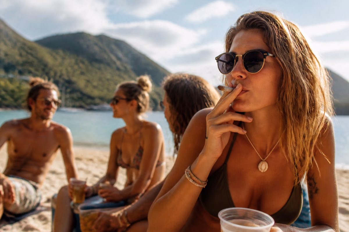 Femme à lunettes de soleil fumant un joint sur une plage tropicale, des amis avec des verres en arrière-plan