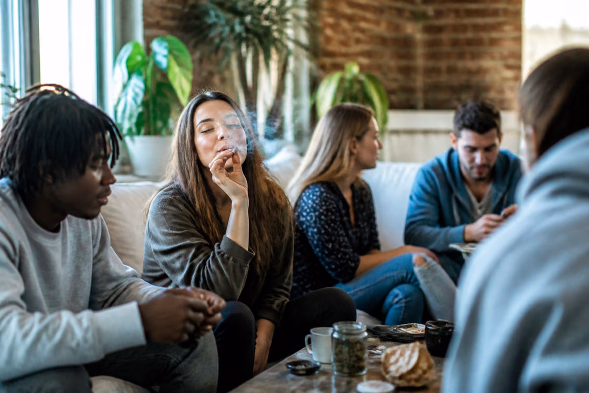 Jeunes adultes sur un canapé, une femme fume un joint, un bocal de cannabis posé sur la table basse