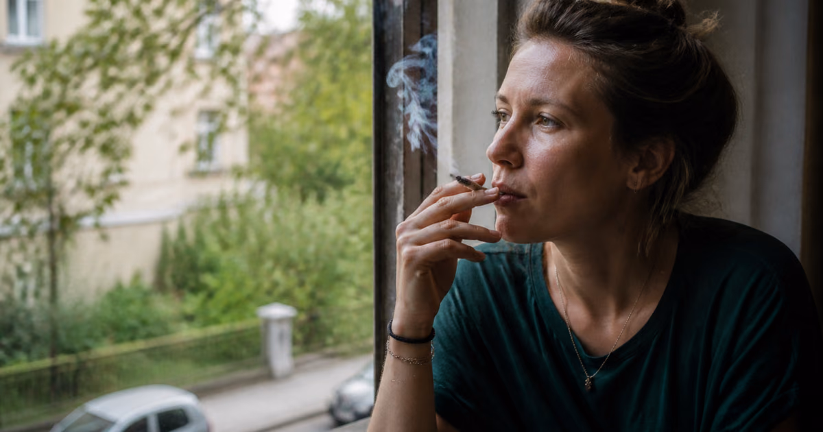 Femme fumant un joint près d’une fenêtre et regardant une rue arborée